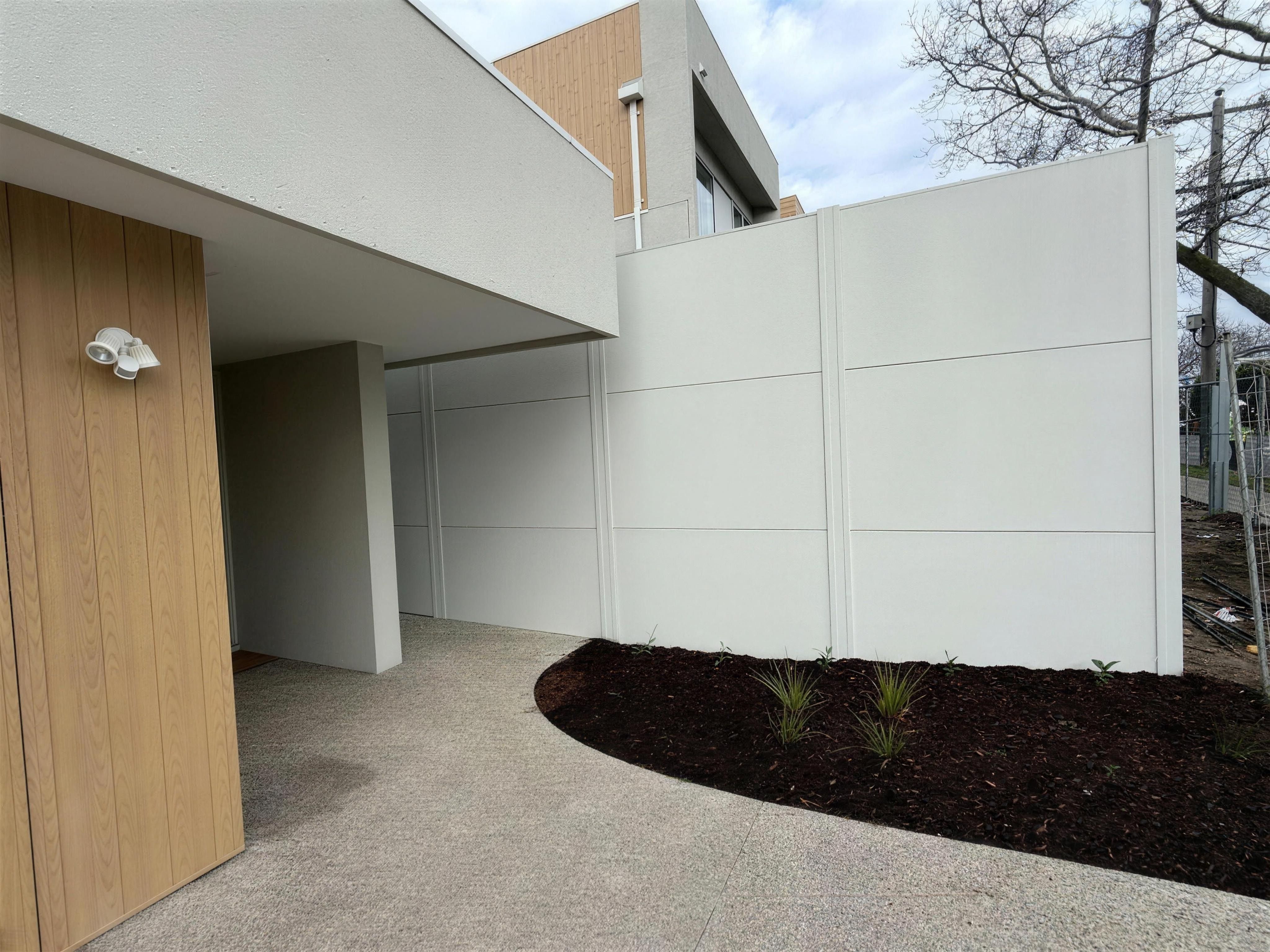 A tall white TrendWall boundary beside a unit entry with a timber-look panel and a new garden bed, construction fencing at the right edge, Brighton.