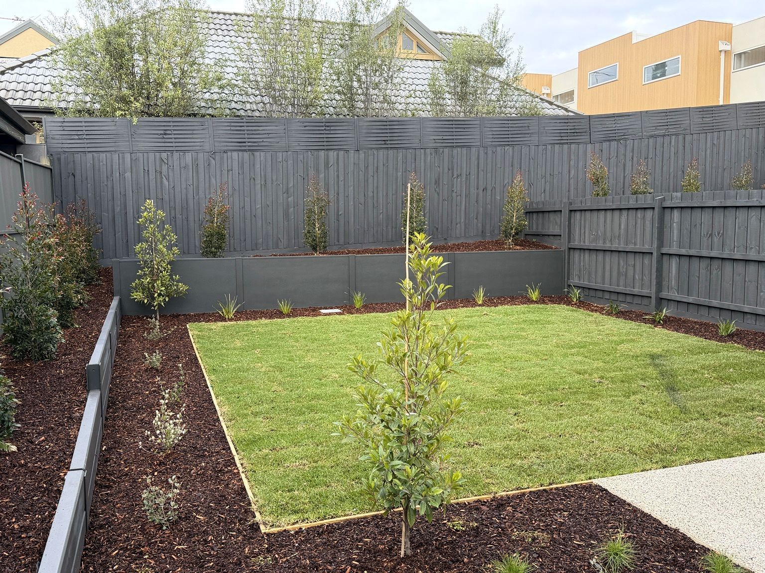 Low charcoal modular retaining walls holding raised garden beds along the back fence of a Brighton unit, with new lawn in front.