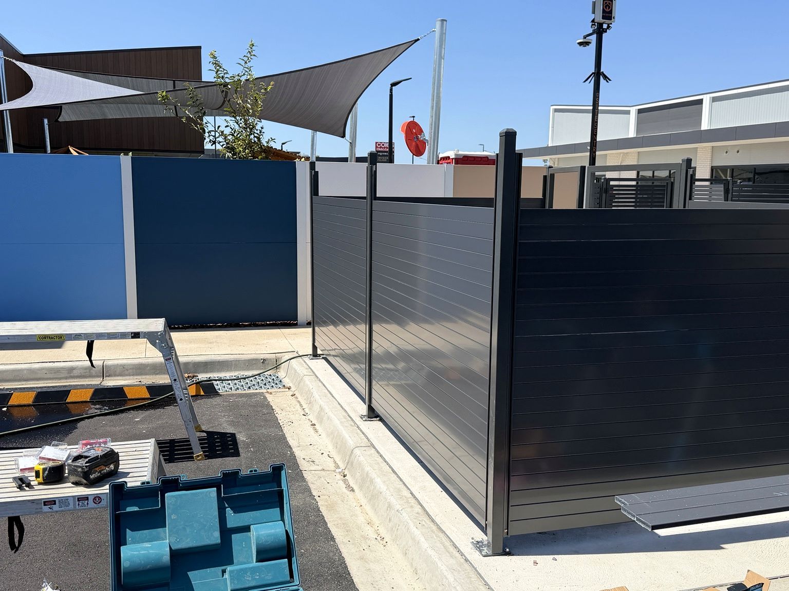 Dark horizontal-slat fence panels going in along a car park kerb at the Craigieburn childcare centre, with a ladder and tools in the foreground.
