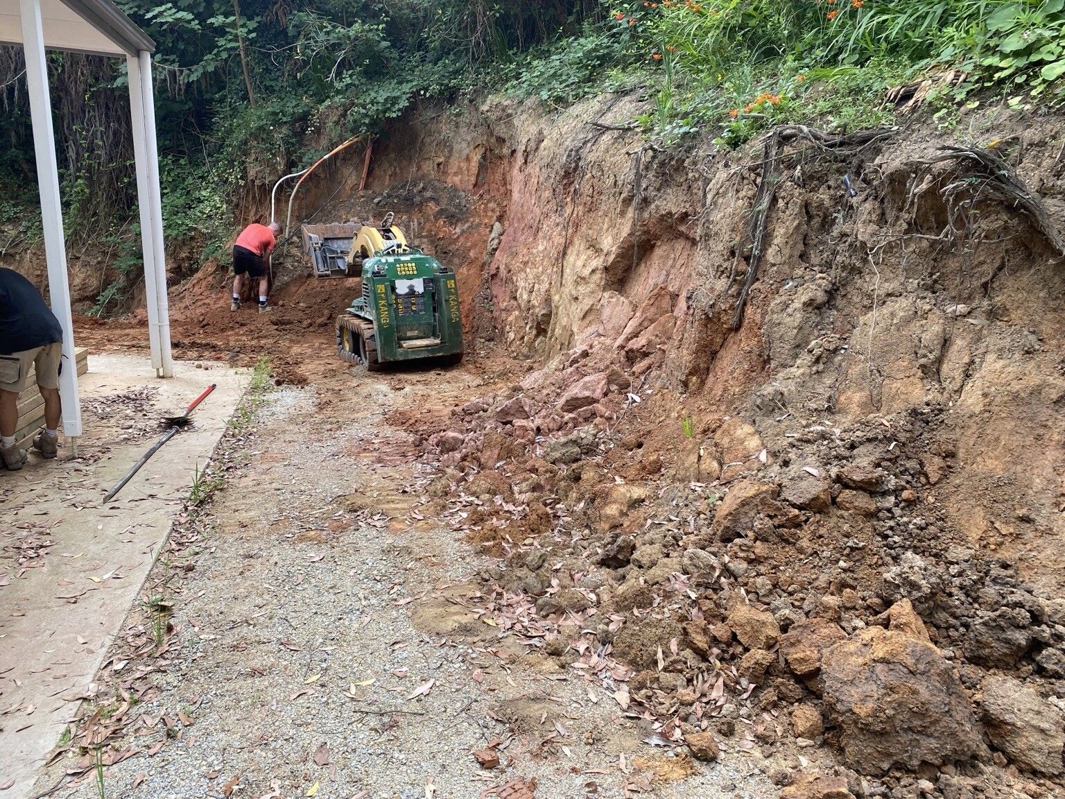 A mini loader excavating a steep red-clay bank beside a house for the Ferny Creek retaining wall.